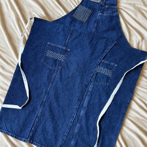 May include: A denim apron with white straps and decorative stitching on the pockets. The apron is made from dark blue denim and features a bib top with a Wrangler label. The apron has two front pockets and a pocket on the bib.