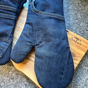 May include: A pair of dark blue denim oven mitts constructed from repurposed jeans fabric. The mitts feature visible pockets, waistbands, and copper-colored stitching details. The fabric has a classic indigo wash with subtle fading and texture.