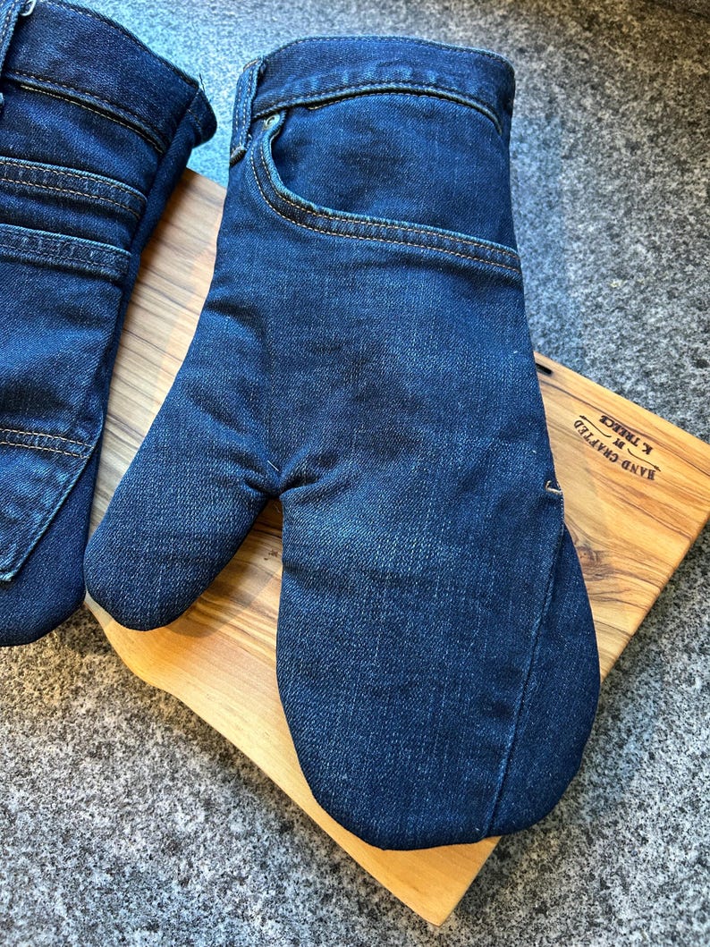 May include: A pair of dark blue denim oven mitts constructed from repurposed jeans fabric. The mitts feature visible pockets, waistbands, and copper-colored stitching details. The fabric has a classic indigo wash with subtle fading and texture.