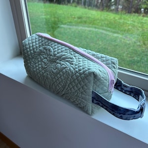 Peut inclure: Une trousse de maquillage matelassée vert sauge avec une fermeture éclair rose et une poignée en jean. Le sac présente un motif en losange et un motif floral décoratif. Le sac rectangulaire est posé sur un rebord de fenêtre blanc.