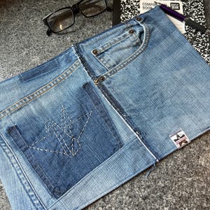 May include: A denim notebook cover made from recycled jeans. The cover features a front pocket, a back pocket with a stitched design, and a small tag. A black composition book, glasses, and a pen are also visible.