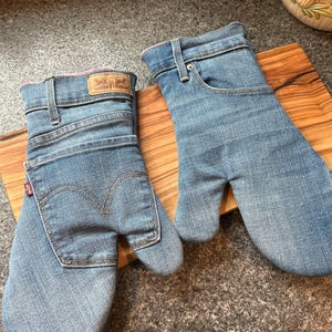 May include: A pair of oven mitts crafted from light blue denim fabric. One mitt features a rear pocket with a red tab and a tan leather patch, while the other displays a front pocket design with a metal rivet and belt loops at the cuff.