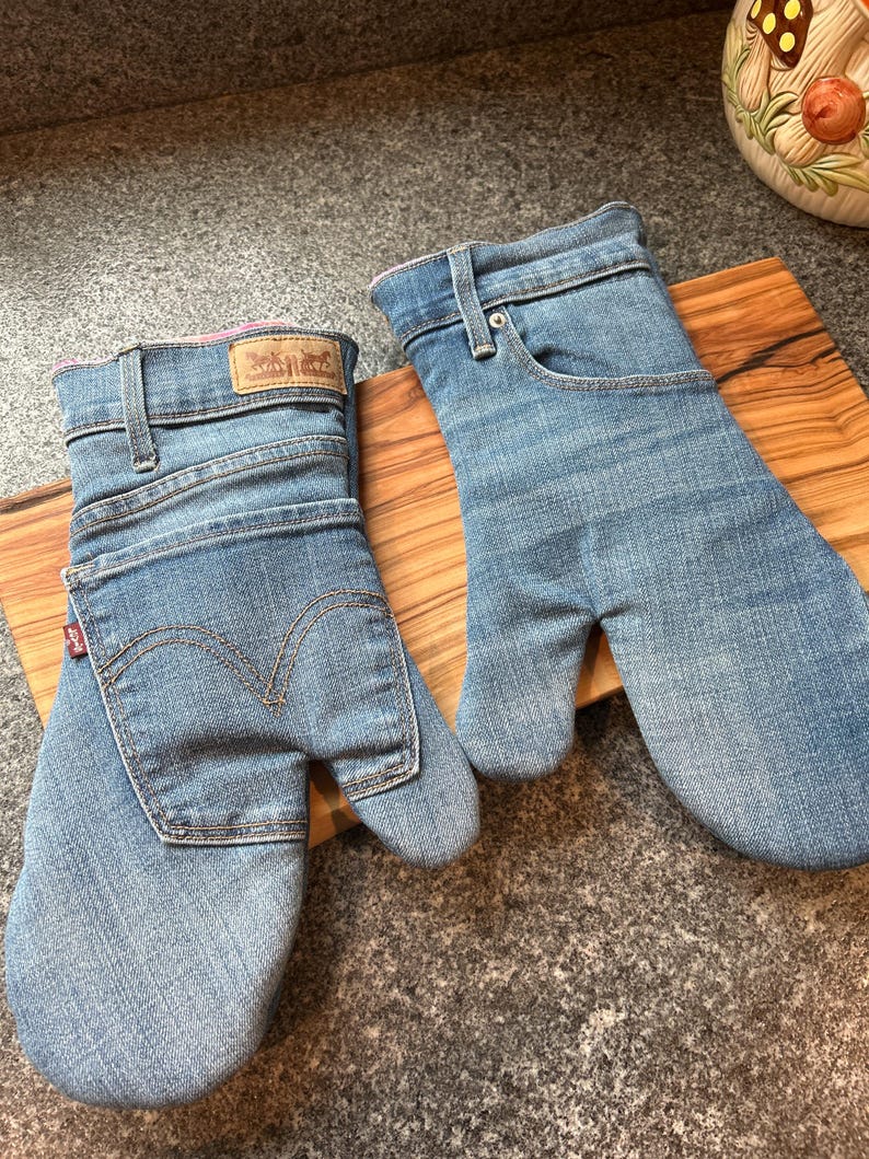 May include: A pair of oven mitts crafted from light blue denim fabric. One mitt features a rear pocket with a red tab and a tan leather patch, while the other displays a front pocket design with a metal rivet and belt loops at the cuff.