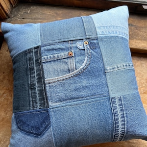 May include: Square denim patchwork pillow in various shades of blue. The pillow features a pocket and other details from repurposed jeans. The pillow is a decorative item for a bed or sofa.