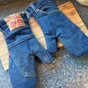 May include: A pair of oven mitts crafted from blue denim fabric. One mitt features a tan Levi Strauss & Co. leather patch with the text W 30 L 30 and a small red tab. The design incorporates denim pocket details, belt loops, and stitching.