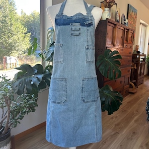 May include: A light blue denim apron featuring a darker blue denim panel at the chest, two upper pockets, and two larger lower pockets. The apron has a halter-style neck strap and is displayed on a white mannequin torso, showcasing a rugged texture.