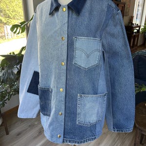 May include: A long-sleeve denim jacket featuring a patchwork design with varying shades of blue. The jacket has a dark blue collar, gold-tone metal snap buttons, and multiple pockets on the front, including a chest pocket with a stitched V-shape.