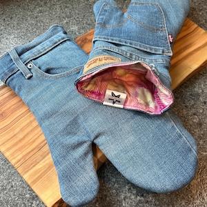 May include: A pair of oven mitts crafted from light blue denim jeans, featuring visible back pockets, belt loops, and a tan leather-style patch. The interior is lined with a pink and white patterned fabric, and a small white label displays a star icon.