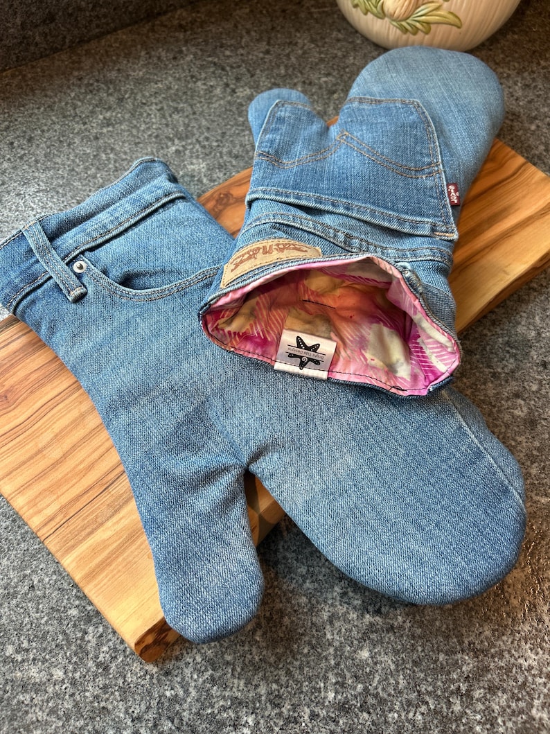 May include: A pair of oven mitts crafted from light blue denim jeans, featuring visible back pockets, belt loops, and a tan leather-style patch. The interior is lined with a pink and white patterned fabric, and a small white label displays a star icon.