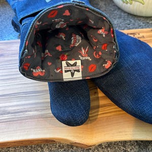 May include: A pair of dark blue denim oven mitts with a gray interior lining featuring a pattern of red lips, hearts, roses, and swallows. A white label inside reads BRIGHT TIDE DESIGNS with a starfish icon. The exterior features a small blue tag.
