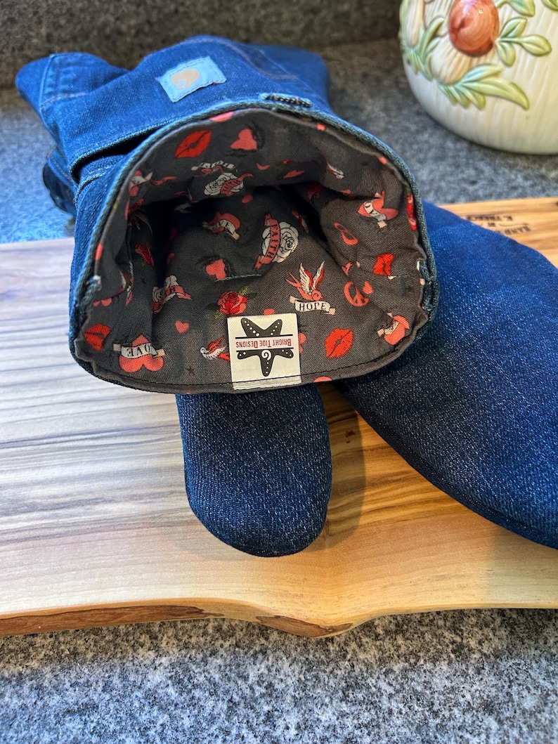 May include: A pair of dark blue denim oven mitts with a gray interior lining featuring a pattern of red lips, hearts, roses, and swallows. A white label inside reads BRIGHT TIDE DESIGNS with a starfish icon. The exterior features a small blue tag.