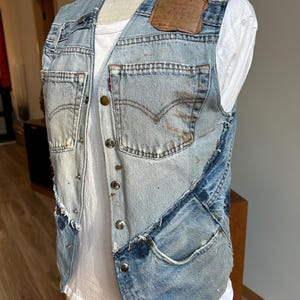 May include: A blue denim vest with a brown leather patch and multiple pockets. The vest is made from repurposed denim and has a distressed look.