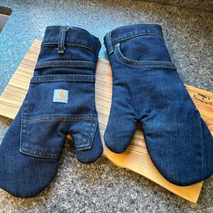 May include: A pair of dark blue denim oven mitts constructed from repurposed jeans. One mitt features a visible Carhartt brand patch and a functional pocket, while the other displays the classic five-pocket jean waistband, belt loops, and pocket.