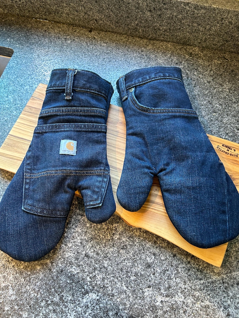 May include: A pair of dark blue denim oven mitts constructed from repurposed jeans. One mitt features a visible Carhartt brand patch and a functional pocket, while the other displays the classic five-pocket jean waistband, belt loops, and pocket.