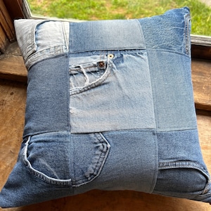 May include: Square denim pillow with a patchwork design. The pillow is made from various shades of blue denim, including light and dark washes. Features include pockets and seams from repurposed jeans.