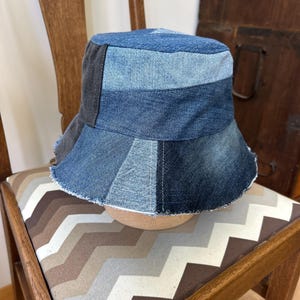 May include: A patchwork denim bucket hat in shades of blue and gray. The hat features a frayed brim and is constructed from various denim pieces. The hat is sitting on a chair with a chevron pattern.