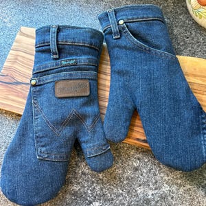 May include: A pair of oven mitts crafted from blue denim fabric, featuring the Wrangler brand logo on a brown leather patch. The mitts include details such as belt loops, a metal rivet, and the signature W stitching pattern on the front pocket.