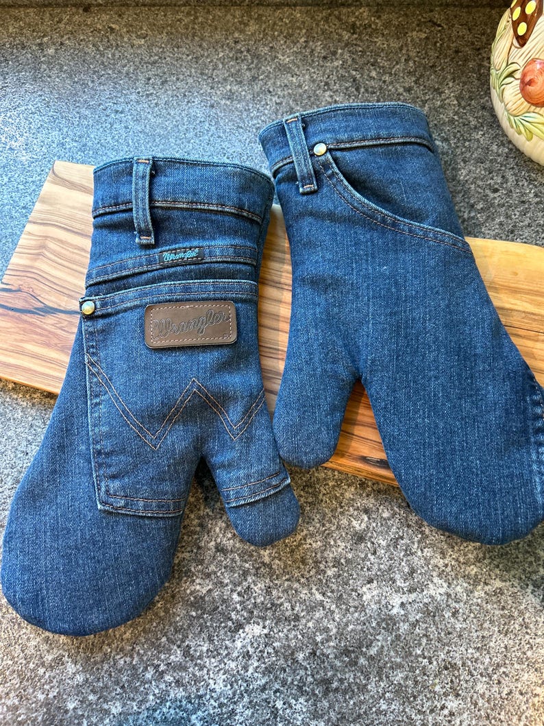 May include: A pair of oven mitts crafted from blue denim fabric, featuring the Wrangler brand logo on a brown leather patch. The mitts include details such as belt loops, a metal rivet, and the signature W stitching pattern on the front pocket.