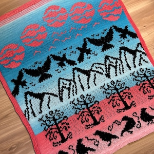 May include: A knitted blanket with a colorful design featuring black birds, trees, and mountains. The blanket has a gradient of blue, pink, and red colors.