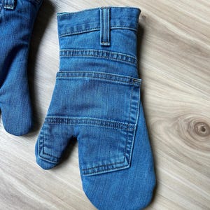 May include: A pair of blue denim oven mitts constructed from repurposed jeans fabric. Each mitt features a waistband with a belt loop at the cuff, visible topstitching, and a square patch pocket on the back of the hand section.