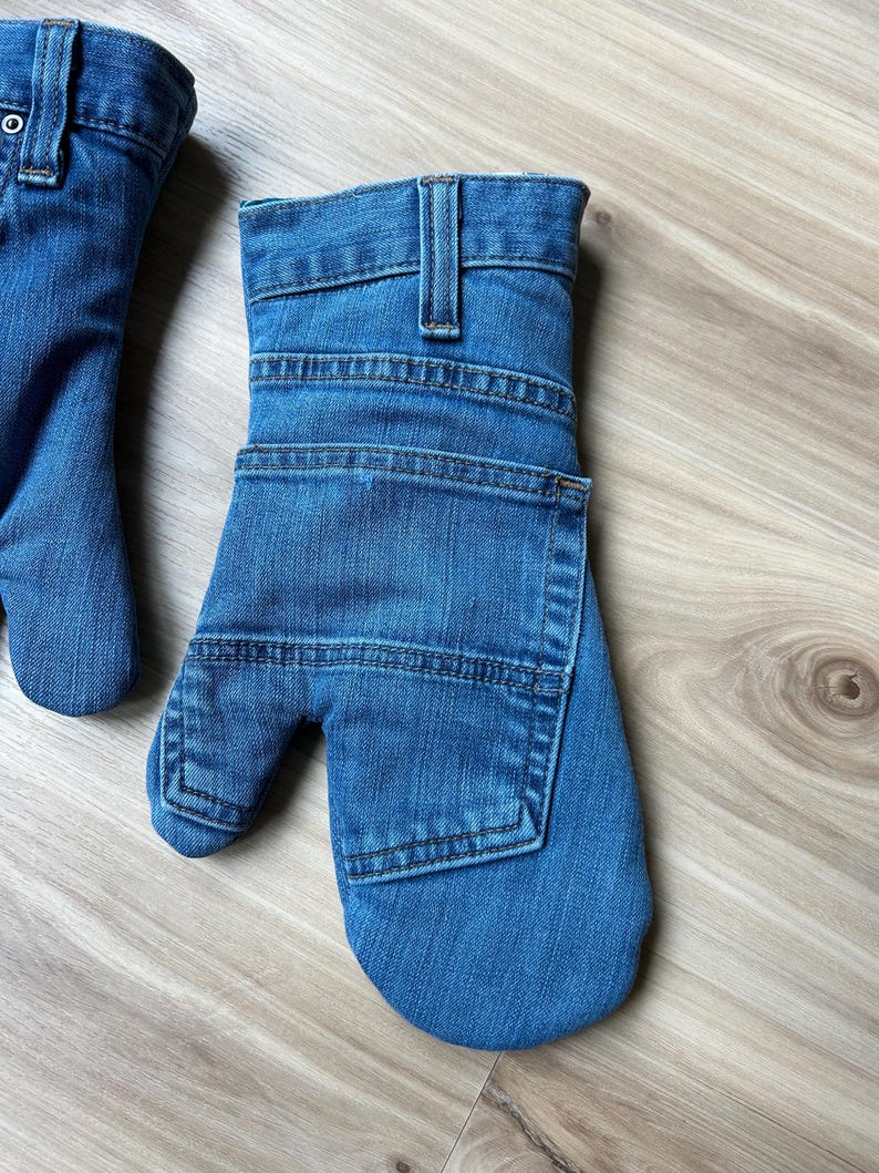 May include: A pair of blue denim oven mitts constructed from repurposed jeans fabric. Each mitt features a waistband with a belt loop at the cuff, visible topstitching, and a square patch pocket on the back of the hand section.