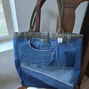 Puede incluir: Un bolso tote de mezclilla con correas y ribetes verde oliva. La bolsa est&aacute; hecha de jeans de mezclilla reciclados, con bolsillos y costuras. La bolsa est&aacute; sobre una silla de madera con un asiento estampado.