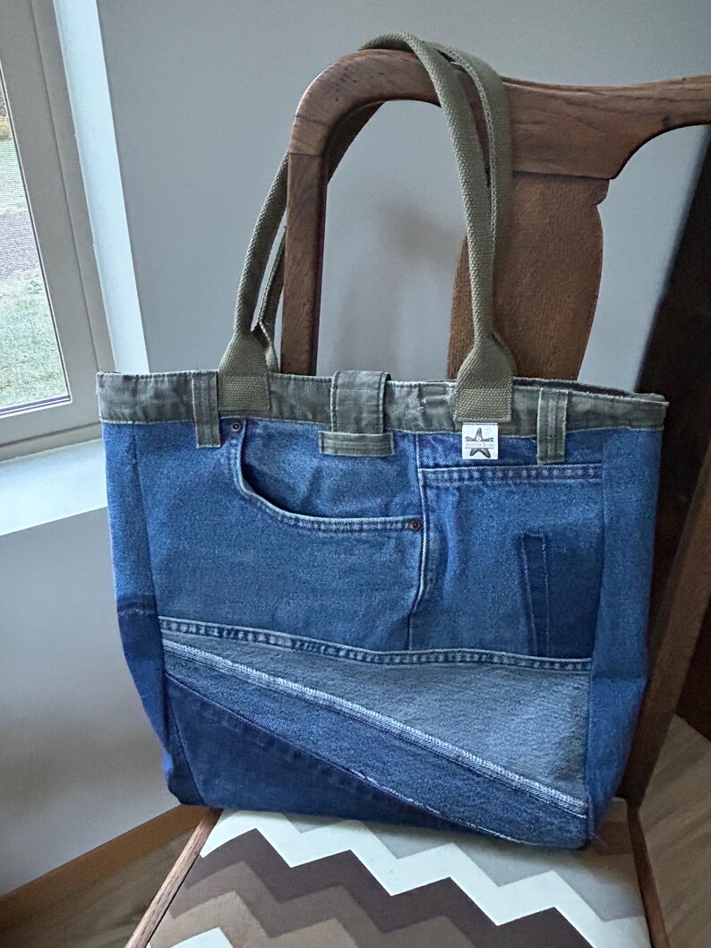 Puede incluir: Un bolso tote de mezclilla con correas y ribetes verde oliva. La bolsa est&aacute; hecha de jeans de mezclilla reciclados, con bolsillos y costuras. La bolsa est&aacute; sobre una silla de madera con un asiento estampado.