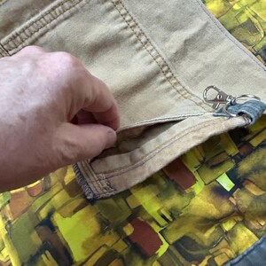 Puede incluir: Primer plano de tela color beige con un bolsillo y costuras visibles. Una mano se mete en el bolsillo. Un cierre plateado est&aacute; adjunto. El fondo presenta un patr&oacute;n abstracto colorido en tonos amarillos, verdes y marrones.