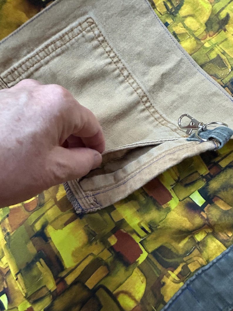 Puede incluir: Primer plano de tela color beige con un bolsillo y costuras visibles. Una mano se mete en el bolsillo. Un cierre plateado est&aacute; adjunto. El fondo presenta un patr&oacute;n abstracto colorido en tonos amarillos, verdes y marrones.