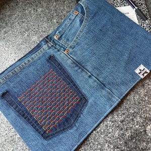 May include: A denim notebook cover with a stitched pocket featuring a red and yellow geometric design. The cover is made from blue denim and has a classic pocket design. A notebook and pen are visible in the background.