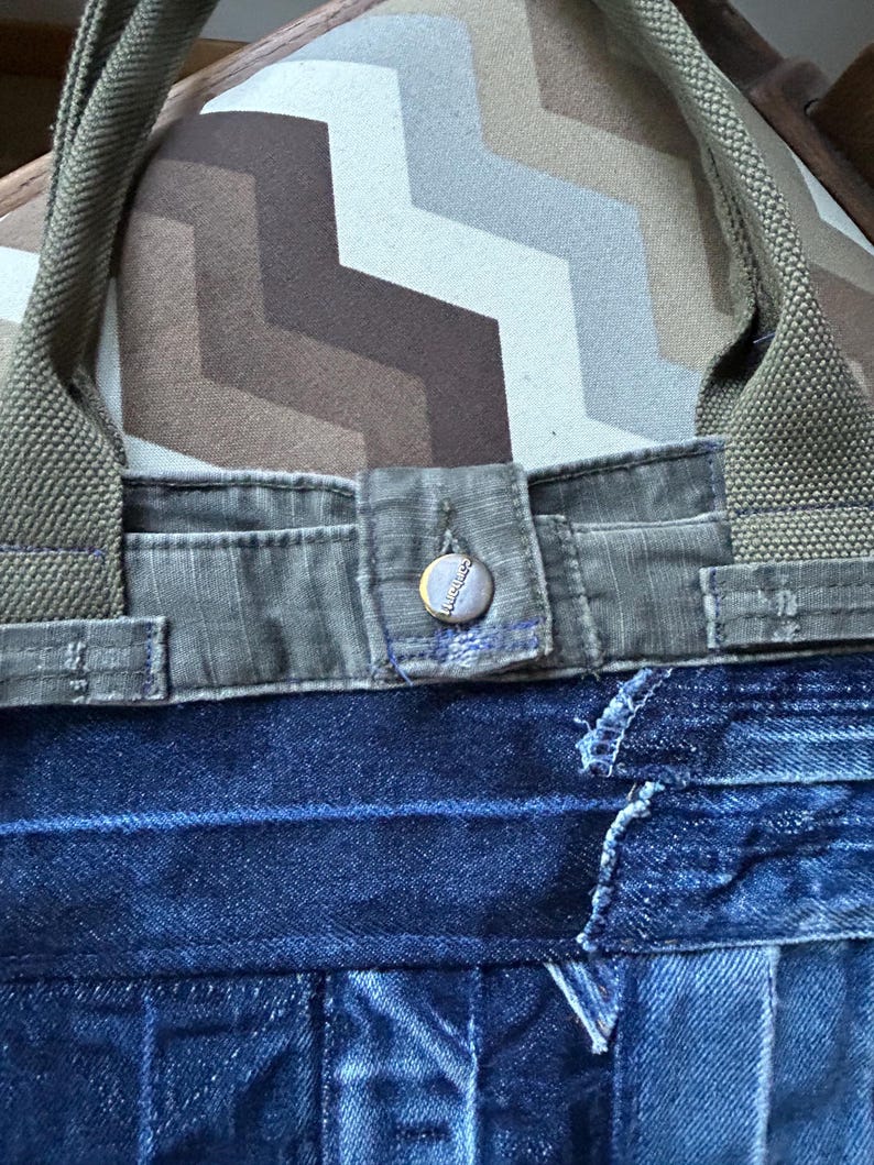 Puede incluir: Un bolso tote de mezclilla con asas y detalles de lona verde oliva. El bolso tiene un cierre de bot&oacute;n y est&aacute; hecho de mezclilla reciclada. El bolso est&aacute; sobre un coj&iacute;n estampado.