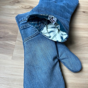 May include: A pair of oven mitts crafted from blue denim fabric, featuring visible pocket stitching and seams. The interior lining is a cream-colored fabric with a dark teal botanical leaf pattern, and a small white brand label is sewn inside.