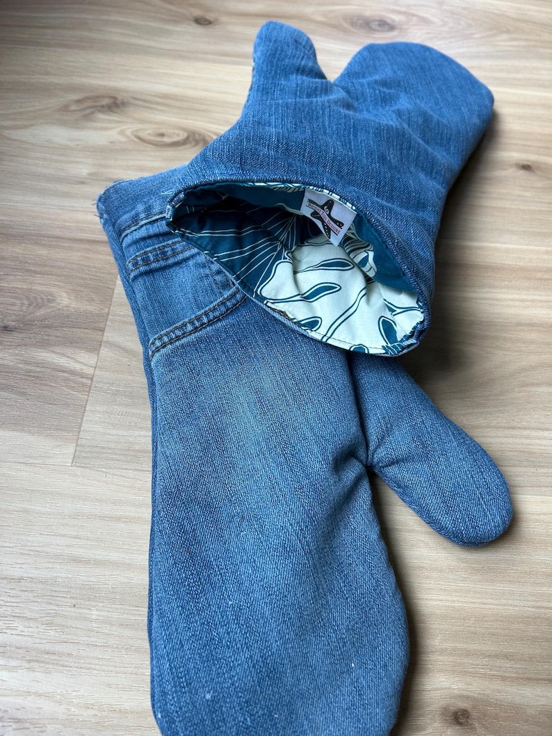May include: A pair of oven mitts crafted from blue denim fabric, featuring visible pocket stitching and seams. The interior lining is a cream-colored fabric with a dark teal botanical leaf pattern, and a small white brand label is sewn inside.