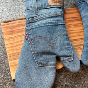 May include: A blue denim oven mitt constructed from repurposed jeans, featuring a rear pocket with signature arcuate stitching, a tan leather-style brand patch at the cuff, and a small red Levi's tab on the side. The interior is lined with pink fabric.