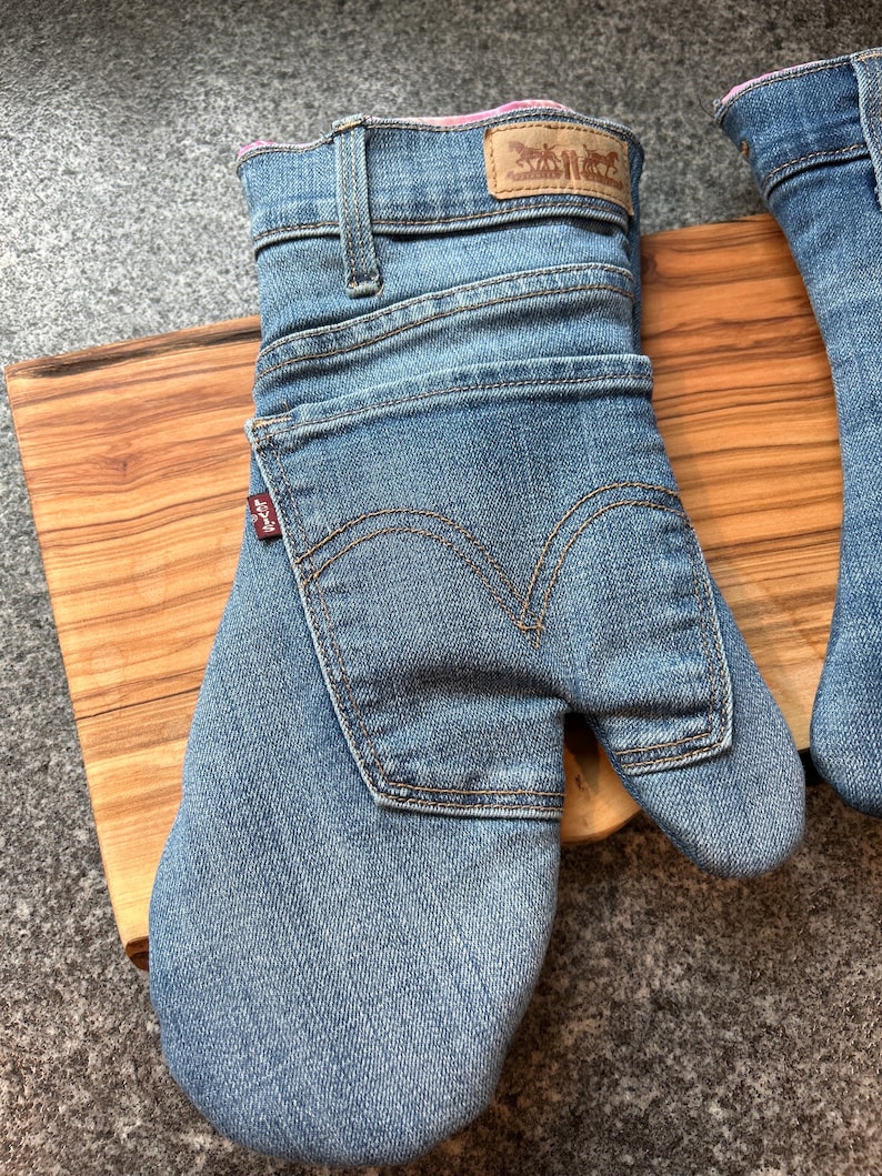 May include: A blue denim oven mitt constructed from repurposed jeans, featuring a rear pocket with signature arcuate stitching, a tan leather-style brand patch at the cuff, and a small red Levi's tab on the side. The interior is lined with pink fabric.