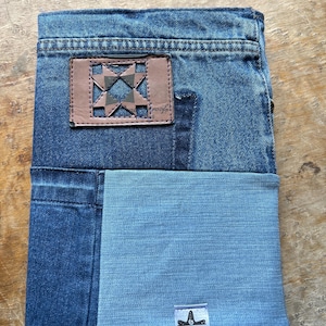 Peut inclure: Une couverture de carnet en jean recyclé. Elle présente un patch en cuir marron avec un motif géométrique et le mot "proof". La partie inférieure est en denim bleu clair. Environ 20 cm de hauteur.