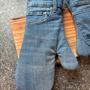 May include: A pair of oven mitts crafted from blue denim fabric, featuring the distinct design of jeans including a waistband, belt loops, a front pocket, and a metal rivet. The interior lining is visible at the cuff, showing a patterned fabric.