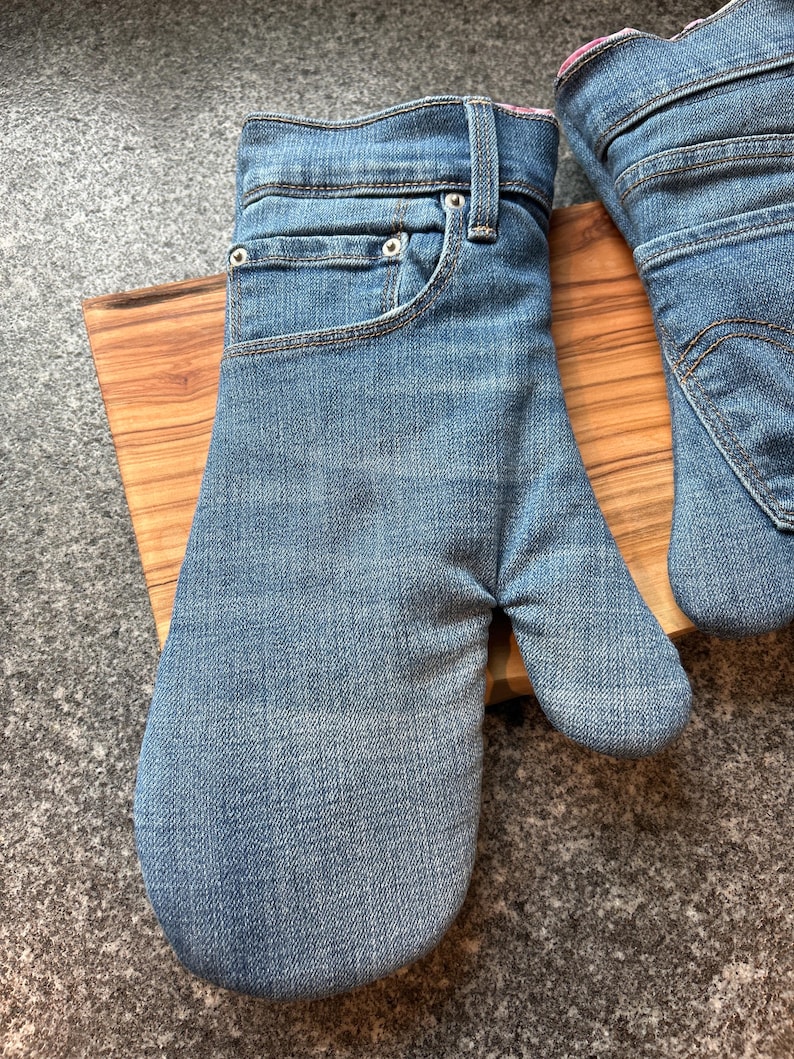 May include: A pair of oven mitts crafted from blue denim fabric, featuring the distinct design of jeans including a waistband, belt loops, a front pocket, and a metal rivet. The interior lining is visible at the cuff, showing a patterned fabric.