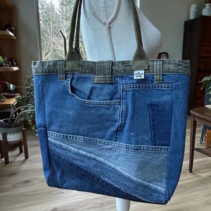 Puede incluir: Un bolso tote de mezclilla azul con detalles en verde oliva. El bolso est&aacute; hecho de mezclilla reciclada y presenta un bolsillo y un parche en forma de estrella. Las asas son de color verde oliva. El bolso se exhibe sobre un soporte blanco.