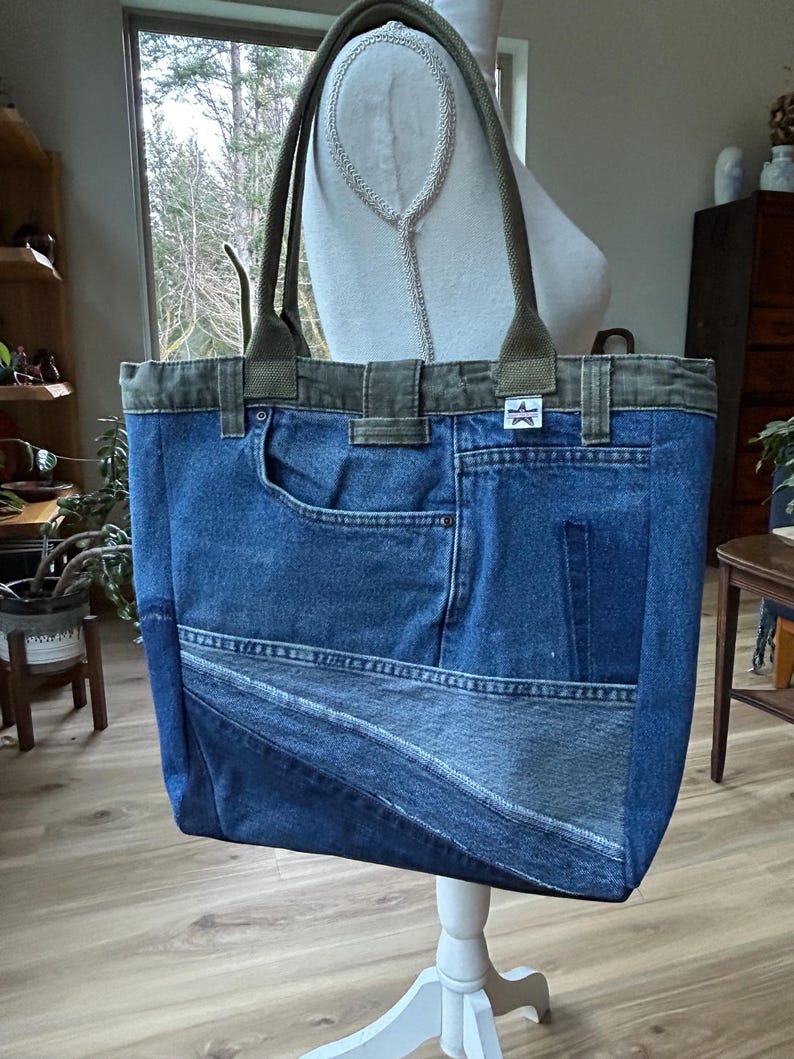 Puede incluir: Un bolso tote de mezclilla azul con detalles en verde oliva. El bolso est&aacute; hecho de mezclilla reciclada y presenta un bolsillo y un parche en forma de estrella. Las asas son de color verde oliva. El bolso se exhibe sobre un soporte blanco.