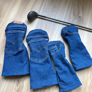 May include: Four blue denim golf club head covers, resembling jean legs, with pockets and stitching details. A golf club is positioned diagonally across the image. The covers are arranged on a light wood-grain surface.