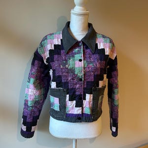 May include: A quilted jacket with a patchwork design in shades of purple, black, pink, and green. The jacket has a gray collar, black buttons, and two front pockets. The jacket is displayed on a white mannequin.