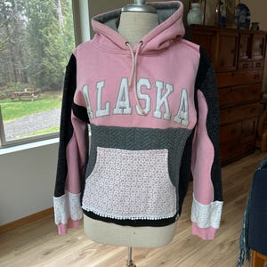 May include: Pink hoodie with the word "ALASKA" in white block letters. The hoodie has a gray hood and a gray and black color block design. The front pocket is made of white lace with a black trim. The sleeves are black and pink.
