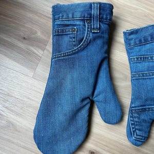 May include: A pair of oven mitts crafted from blue denim fabric, designed to resemble the upper portion of a pair of jeans. Each mitt features a functional pocket, a metal rivet, a belt loop, and visible stitching details consistent with denim pants.