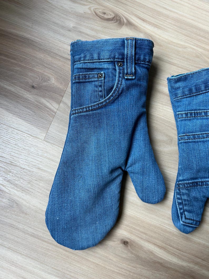 May include: A pair of oven mitts crafted from blue denim fabric, designed to resemble the upper portion of a pair of jeans. Each mitt features a functional pocket, a metal rivet, a belt loop, and visible stitching details consistent with denim pants.