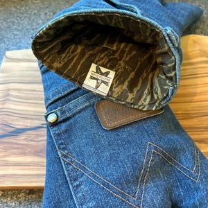 May include: A blue denim oven mitt featuring a brown leather patch with the Wrangler logo and a gold-tone metal rivet. The interior is lined with a brown and tan camouflage fabric, and a white tag displays the text Bright Tide Designs.