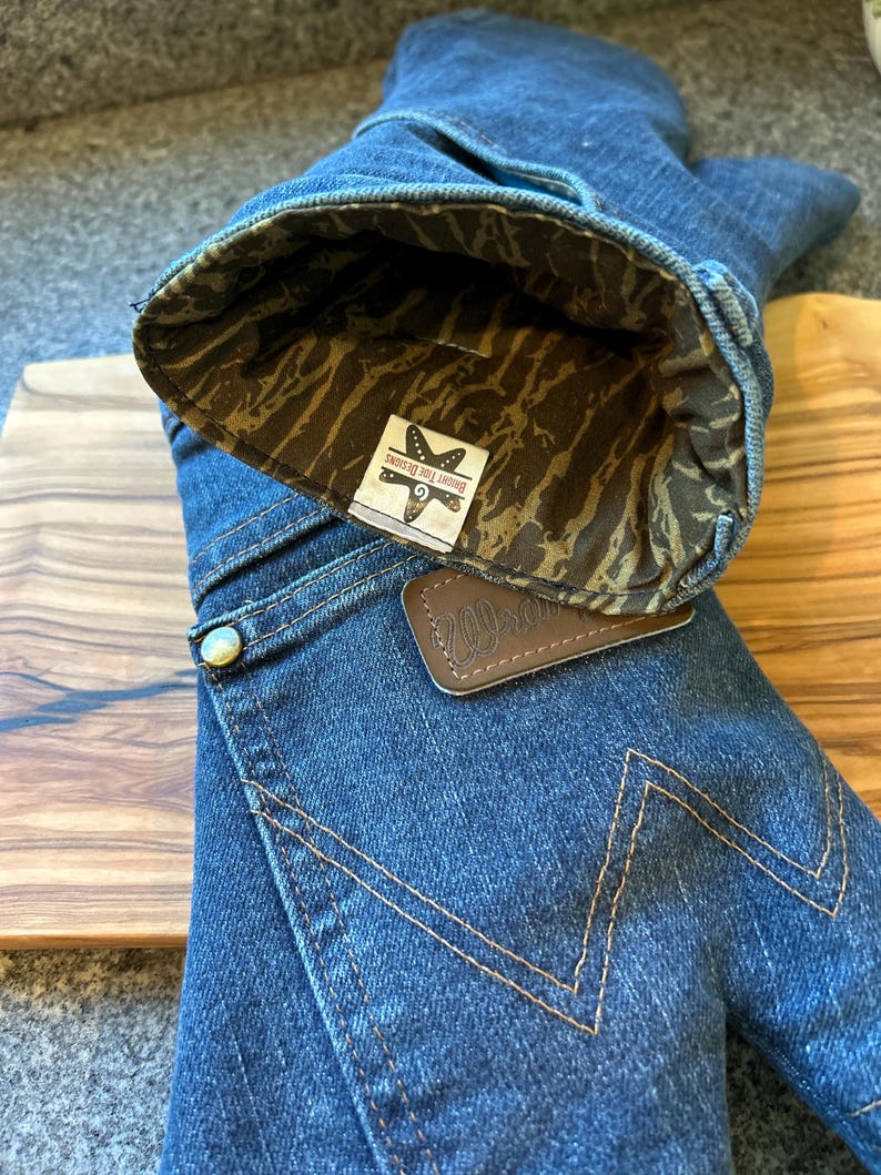May include: A blue denim oven mitt featuring a brown leather patch with the Wrangler logo and a gold-tone metal rivet. The interior is lined with a brown and tan camouflage fabric, and a white tag displays the text Bright Tide Designs.