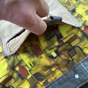 Puede incluir: Primer plano de un bolsillo de tela beige con una costura azul, que revela un forro con estampado abstracto vibrante en amarillo, marr&oacute;n y verde. El bolsillo se est&aacute; abriendo, mostrando el dise&ntilde;o interior.