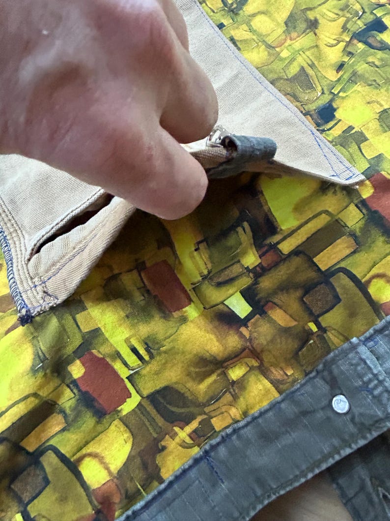 Puede incluir: Primer plano de un bolsillo de tela beige con una costura azul, que revela un forro con estampado abstracto vibrante en amarillo, marr&oacute;n y verde. El bolsillo se est&aacute; abriendo, mostrando el dise&ntilde;o interior.