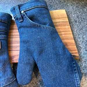 May include: A pair of oven mitts crafted from blue denim fabric, featuring classic jean details such as a curved pocket opening, a metal rivet, and a belt loop at the wrist. The mitts have visible topstitching and a rounded thumb section.